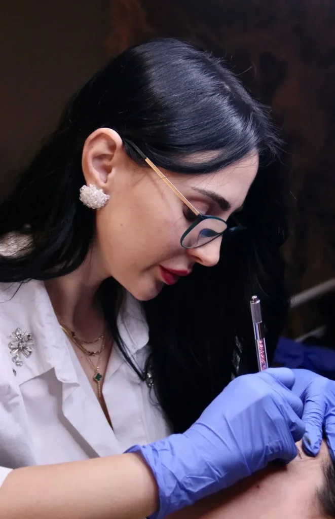 Martina Cetaj in glasses and gloves, drawing a brow design on a client