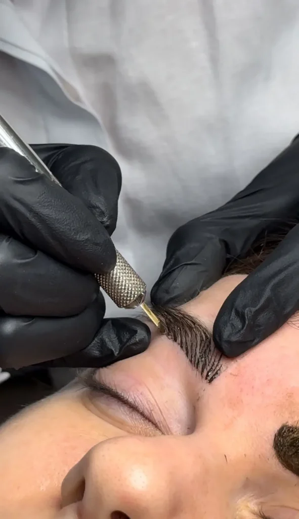 Gloved hands using a microblading tool to draw hair strokes on a brow