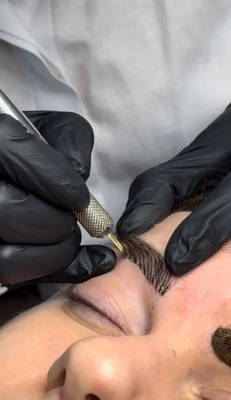 Gloved hands using a microblading tool to draw hair strokes on a brow